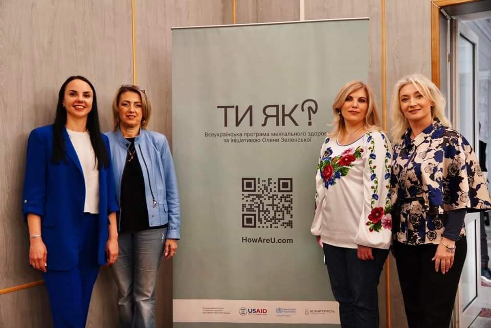 Чотири жінки стоять поруч з банером, на якому написано "Ти як?".
