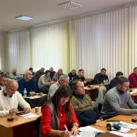 В залі  багато людей сидять за столами, перед ними записники 